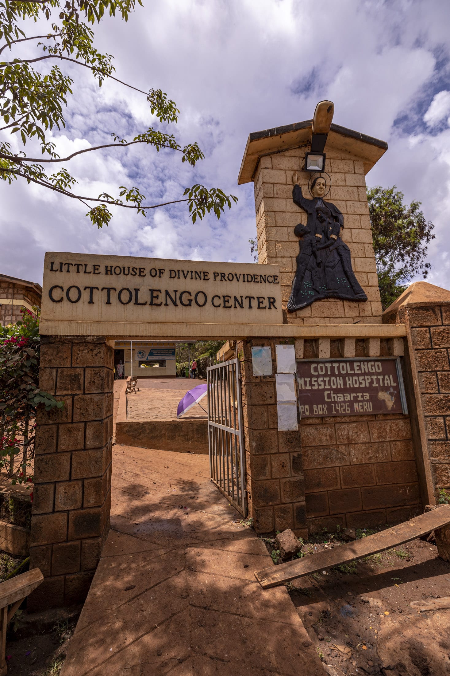 Eingang zum Cottolengo Center mit einem Schild mit der Aufschrift „Kleines Haus der göttlichen Vorsehung – Cottolengo Center“. Über dem Schild steht auf einer Steinsäule eine Statue. Es gibt ein Tor und ein zweites Schild mit der Adresse und den Einzelheiten des Cottolengo Mission Hospital. © Fotografie Tomas Rodriguez