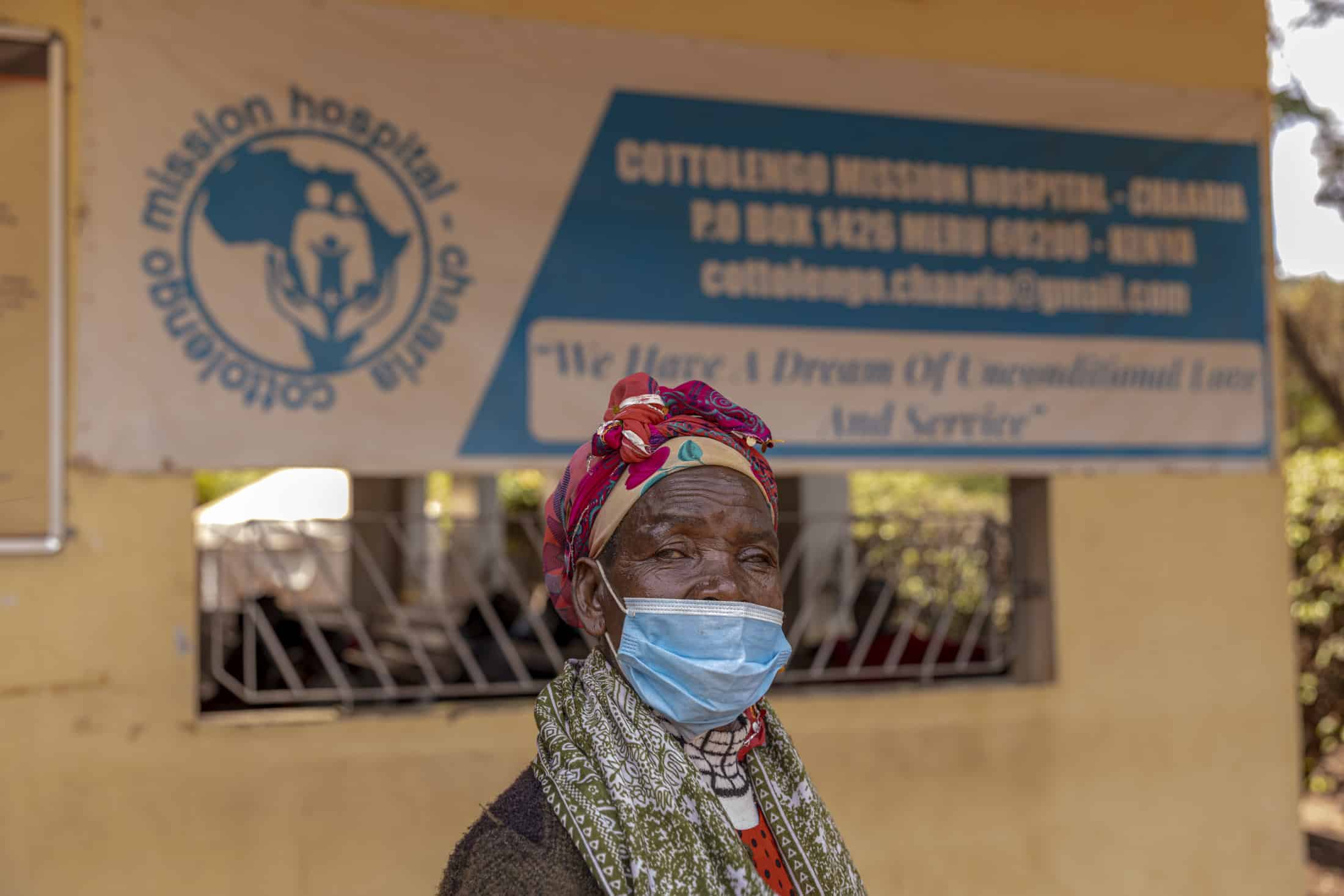 Eine ältere Frau mit Gesichtsmaske steht vor einem Gebäude des Cotolengo Mission Hospital in Chaaria, Kenia. Sie trägt farbenfrohe Kleidung und ein Kopftuch. Im Hintergrund ist ein Banner mit dem Namen und den Kontaktdaten des Krankenhauses zu sehen. © Fotografie Tomas Rodriguez