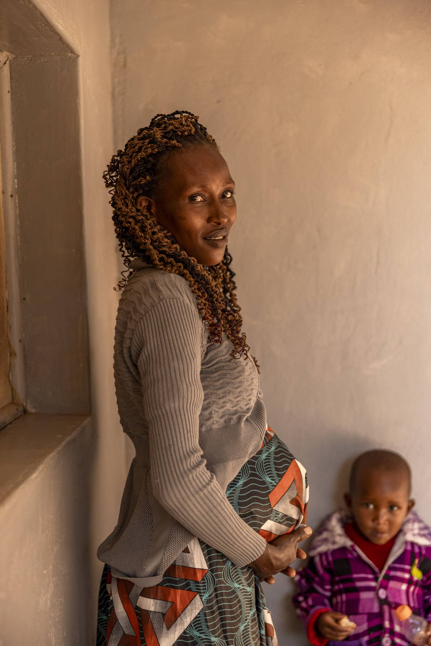 Eine schwangere Frau mit geflochtenen Haaren, die ein graues langärmliges Oberteil und einen gemusterten Rock trägt, steht drinnen neben einem Fenster. Sie legt ihre Hände auf ihren Bauch, während im Hintergrund, teilweise unscharf, ein kleines Kind in einer karierten Jacke steht. © Fotografie Tomas Rodriguez