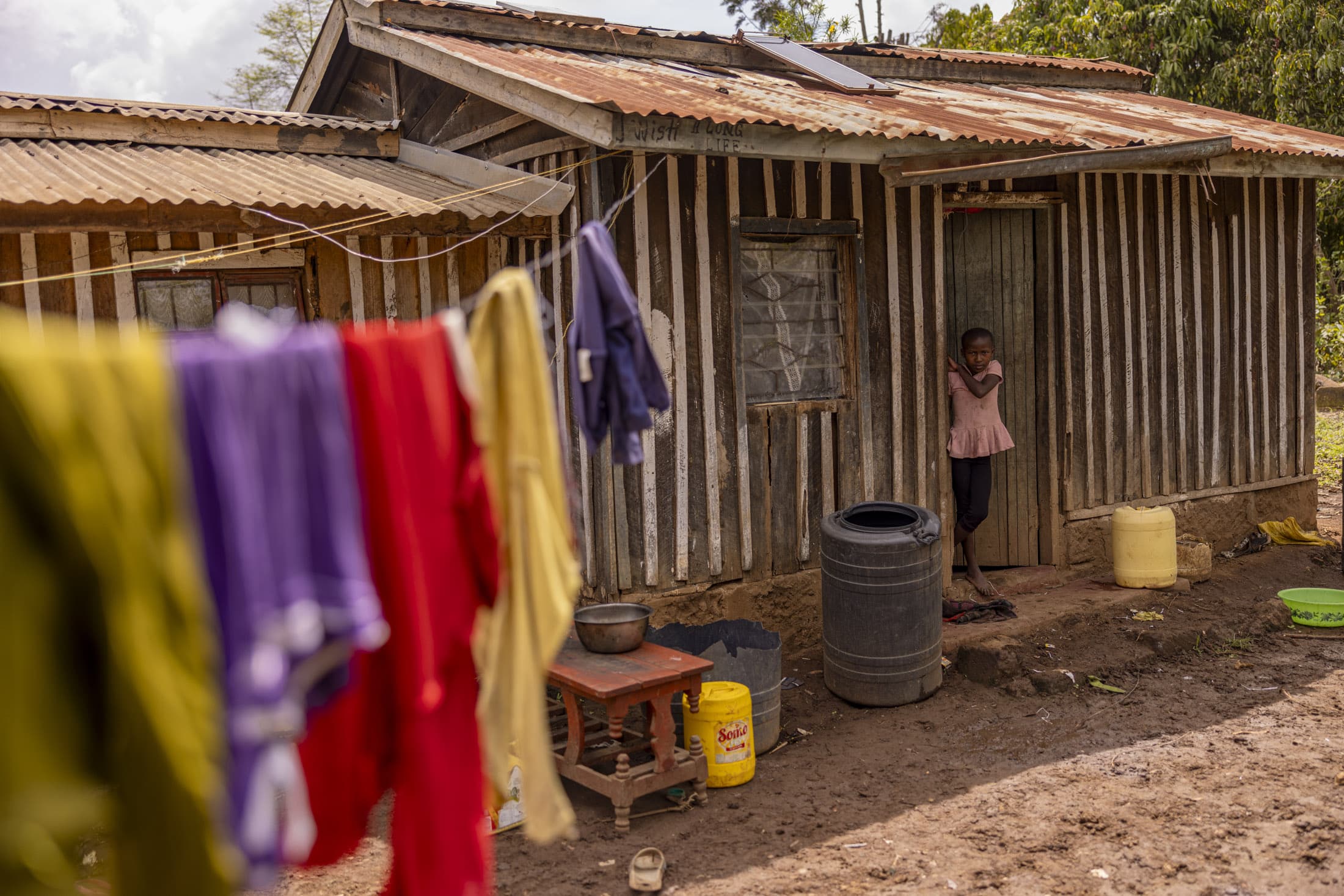 Ein Kind steht in der Tür eines kleinen, rustikalen Hauses mit Wellblechdach und vertikalen Holzlatten. Im Vordergrund hängen bunte Kleidungsstücke auf einer Leine, während im Hof Haushaltsgegenstände und Wasserbehälter verstreut sind. © Fotografie Tomas Rodriguez