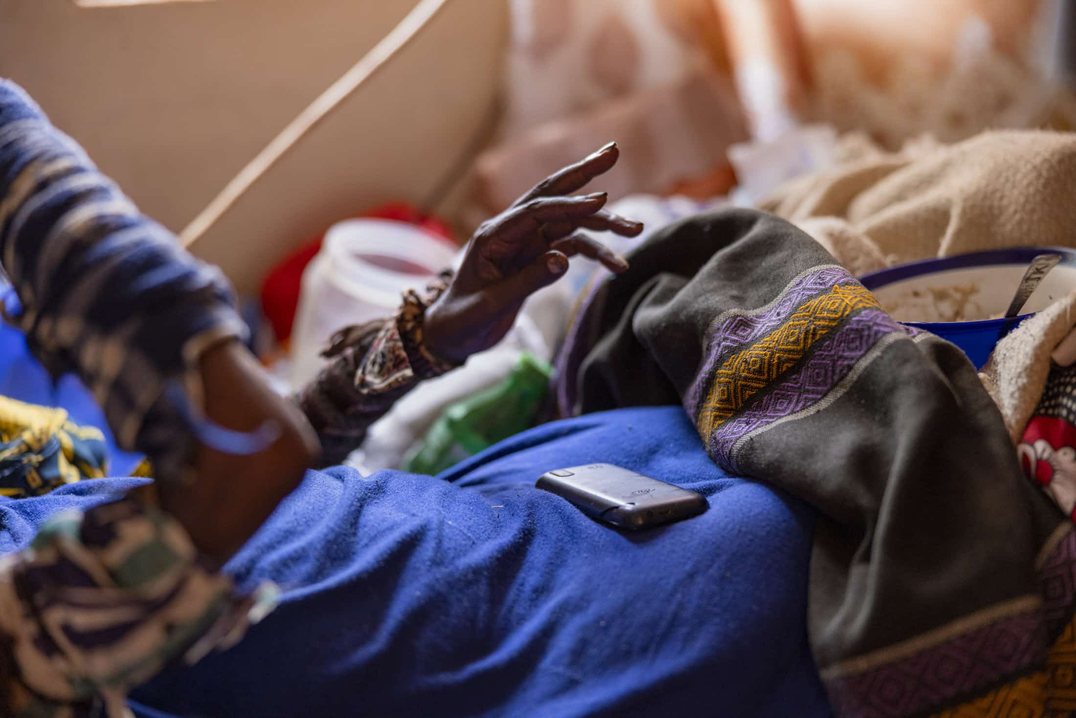 Die Hand einer Person streckt sich aus und ruht auf einer Oberfläche, die mit einer bunten Decke bedeckt ist. Auf der Decke liegt ein Smartphone, im Hintergrund sind verschiedene Haushaltsgegenstände zu sehen, die eine gemütliche, überladene Atmosphäre schaffen. © Fotografie Tomas Rodriguez