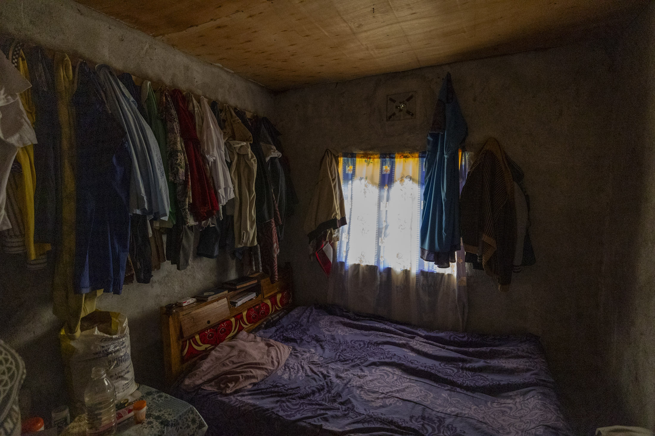 Ein kleiner, schwach beleuchteter Raum mit niedriger Holzdecke. An den Betonwänden hängen Kleider. Ein Bett mit einer lila Decke steht neben einem Fenster mit gemusterten Vorhängen. Unter dem Fenster befindet sich ein Regal mit verschiedenen Gegenständen. Licht fällt durch die Vorhänge und erhellt einen Teil des Raumes. © Fotografie Tomas Rodriguez