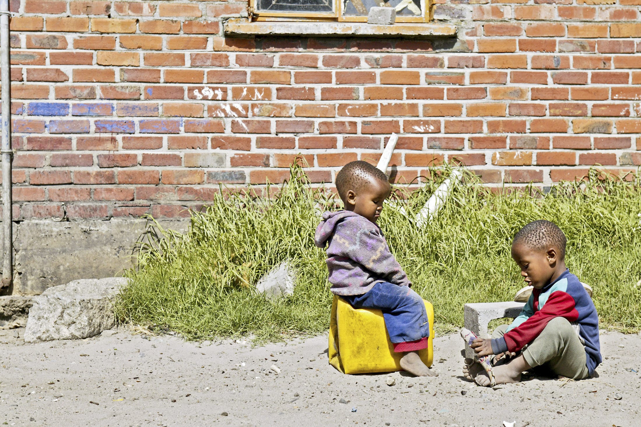 Zwei kleine Kinder spielen draußen vor einer Backsteinmauer mit einem kleinen Fenster. Ein Kind sitzt auf einem gelben Container, während das andere auf dem Boden sitzt. Sie sind von Gras und Schutt umgeben und spielen miteinander. © Fotografie Tomas Rodriguez