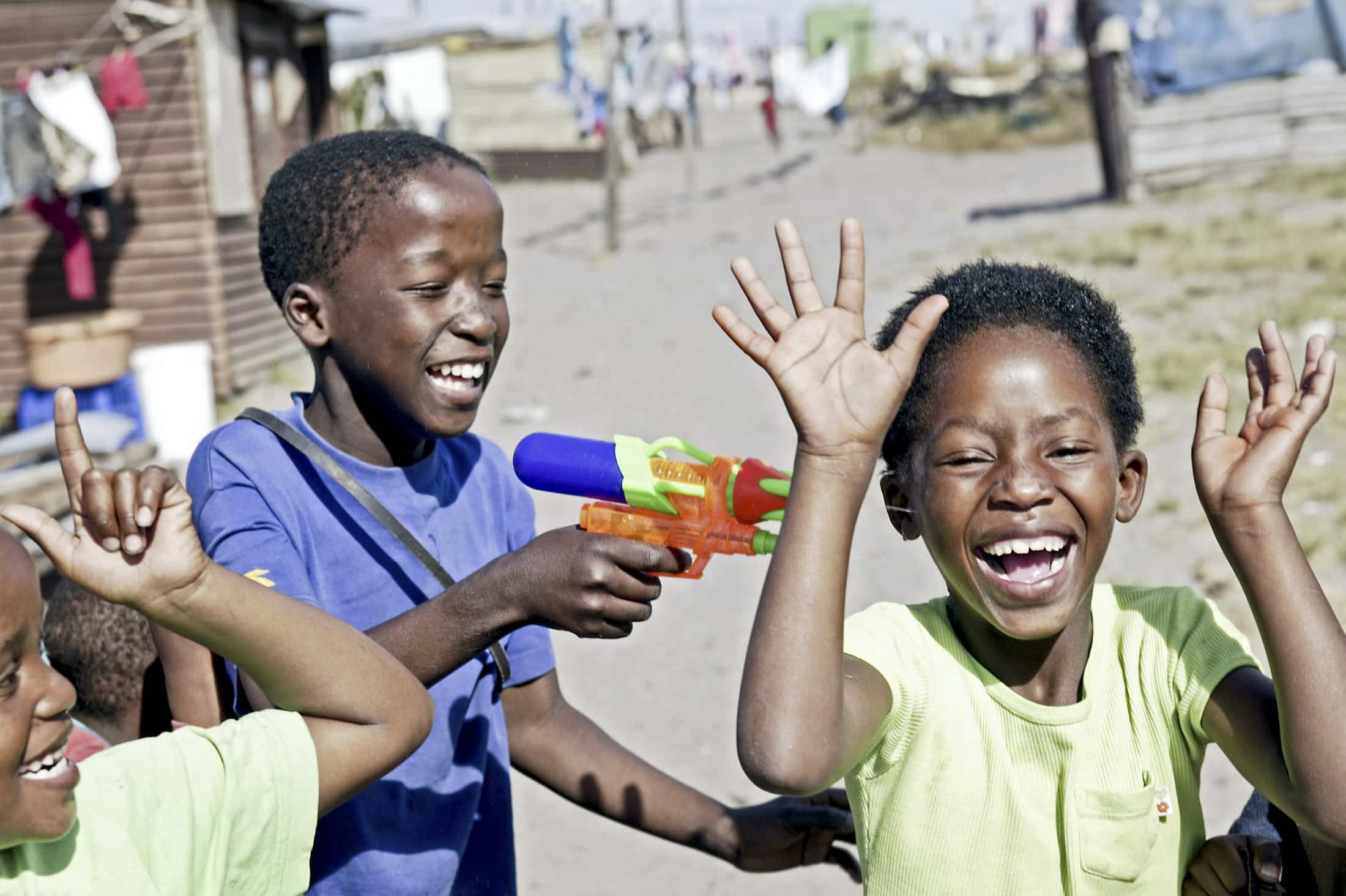 Eine Gruppe lächelnder Kinder im Freien. Ein Junge zielt spielerisch mit einer bunten Wasserpistole auf einen anderen Jungen, der lachend die Hände hochhält. Sie haben Spaß in der Sonne in einer trockenen und staubigen Gegend mit kleinen Häusern und hängenden Kleidern im Hintergrund. © Fotografie Tomas Rodriguez