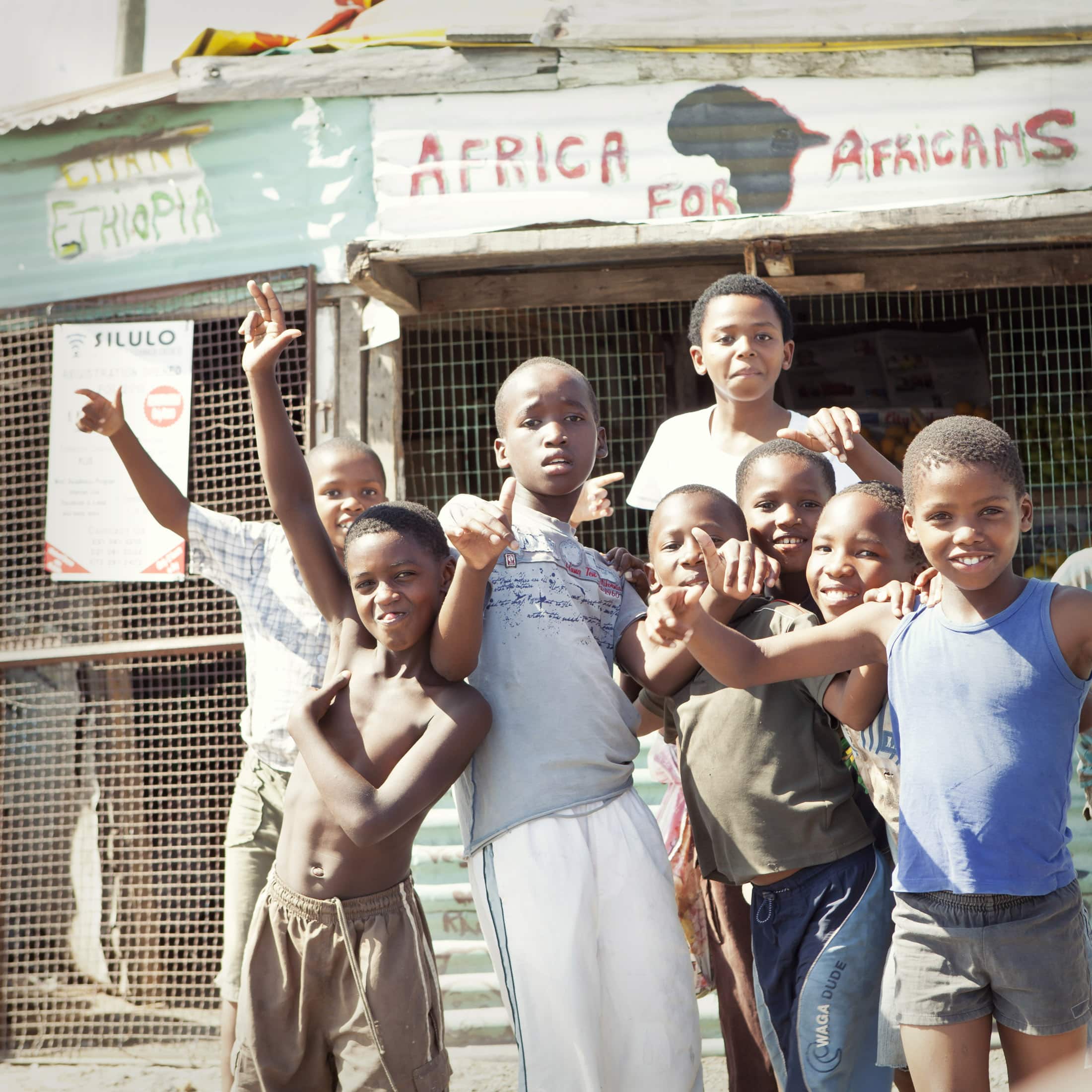 Eine Gruppe lebhafter Kinder lächelt und posiert vor einem Gebäude, auf dem Botschaften wie „Afrika für Afrikaner“ und „Äthiopien“ stehen, für die Kamera. Die Szene ist herzlich und lebhaft, und die Kinder zeigen verschiedene spielerische Gesten. © Fotografie Tomas Rodriguez