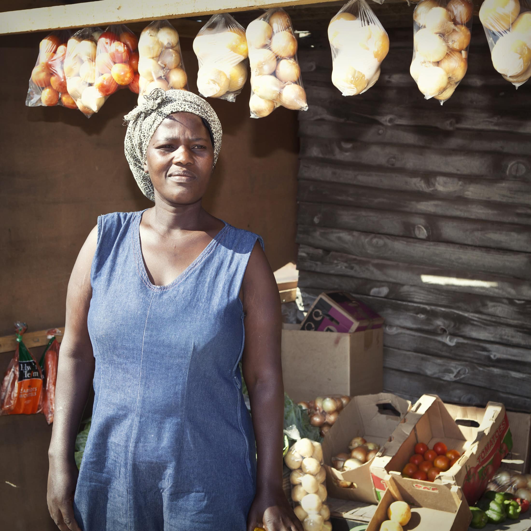 Eine Frau in einem blauen ärmellosen Kleid und Kopftuch steht vor einem Marktstand. Über dem Stand hängen Säcke mit Zwiebeln und Tomaten, darunter sind in Kisten verschiedene Produkte, darunter Tomaten und Kartoffeln, angeordnet. Der Holzstand hat eine rustikale Atmosphäre. © Fotografie Tomas Rodriguez