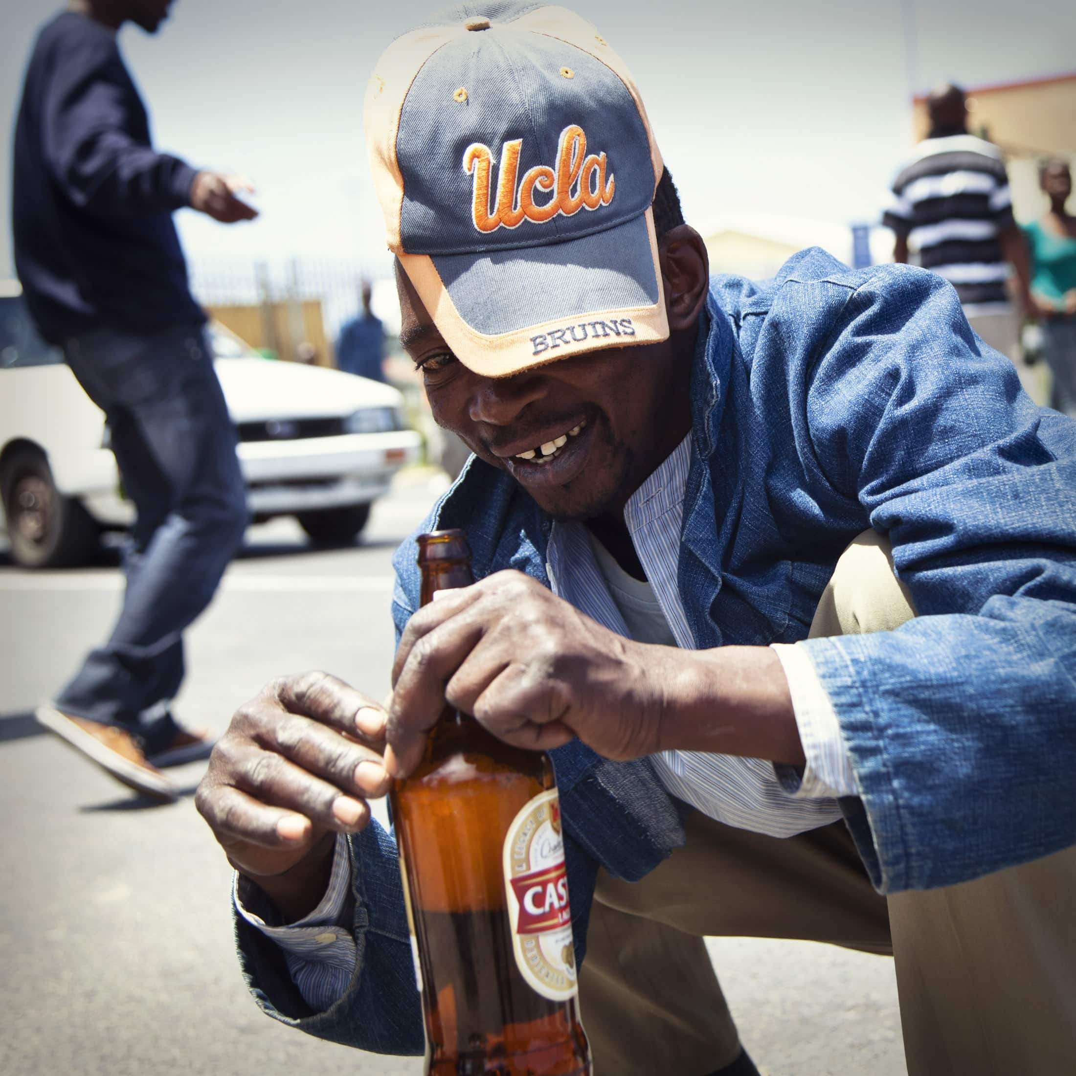 Ein Mann mit einer UCLA-Baseballkappe und einer Jeansjacke hockt lächelnd auf der Straße und hält eine Flasche Castle Lagerbier in der Hand. Im Hintergrund sind weitere Personen und ein weißes Auto zu sehen, darüber ein klarer, sonniger Himmel. © Fotografie Tomas Rodriguez