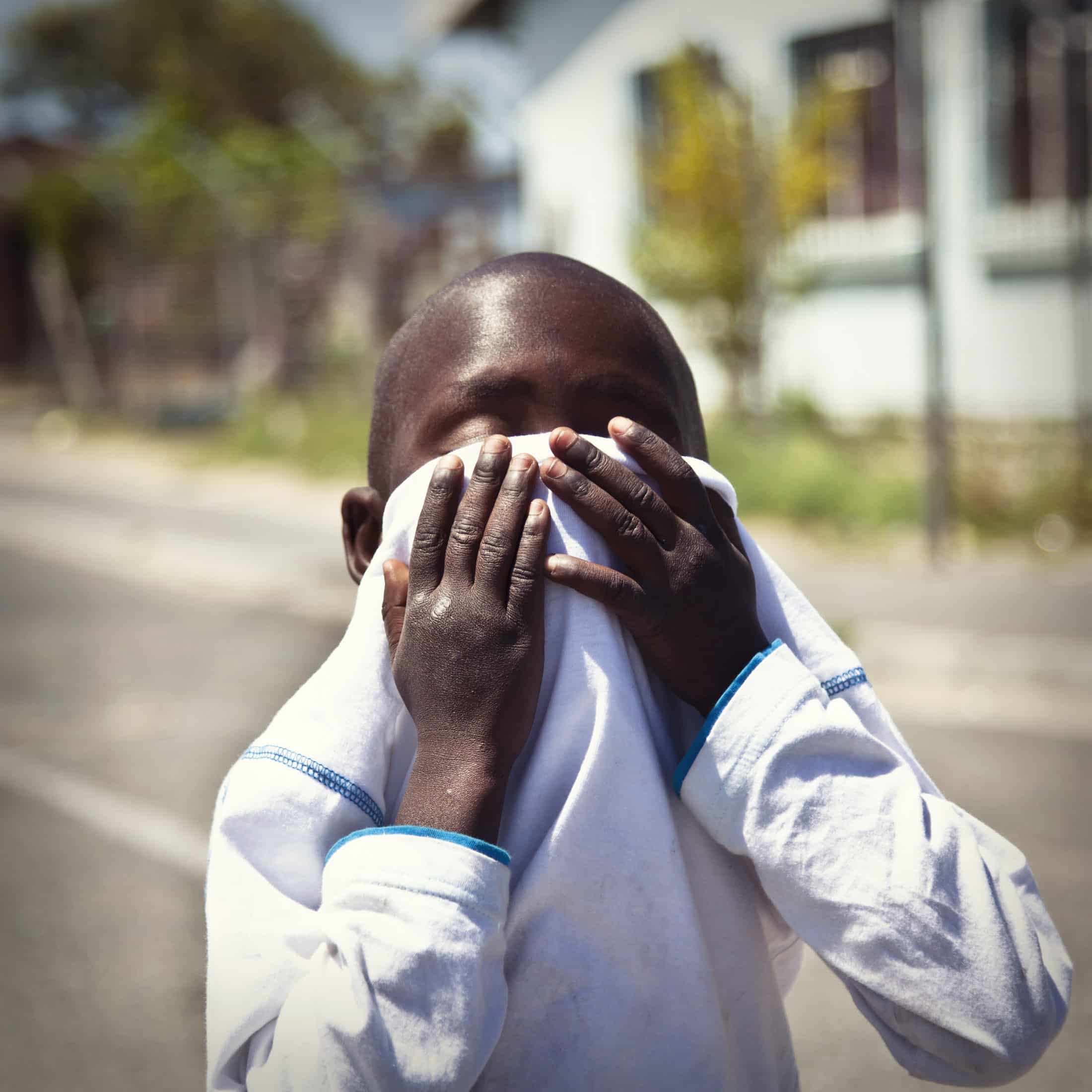 Ein kleiner Junge steht im Freien und verdeckt sein Gesicht mit seinem weißen Langarmshirt. Im Hintergrund sind verschwommene Häuser und Bäume an einem sonnigen Tag zu sehen. © Fotografie Tomas Rodriguez