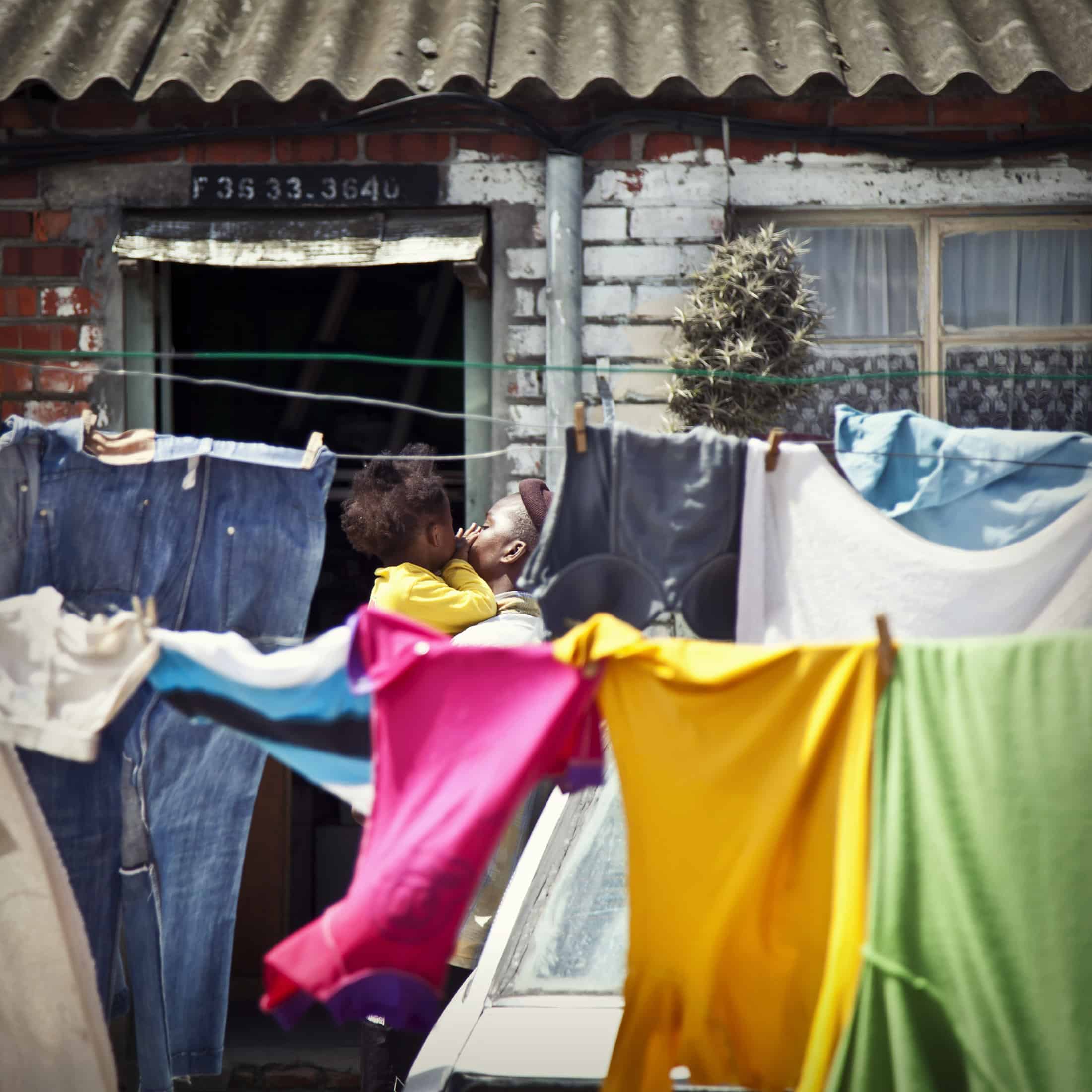 Eine Person, die ein Kind hält, steht vor einem Backsteinhaus mit Wellblechdach. Die Person ist teilweise durch bunte Kleidung verdeckt, die auf einer Wäscheleine hängt, darunter Jeans, ein rotes Hemd und ein gelbes Hemd. Das Haus hat eine grüne Tür und ein Fenster mit einem weißen Vorhang. © Fotografie Tomas Rodriguez