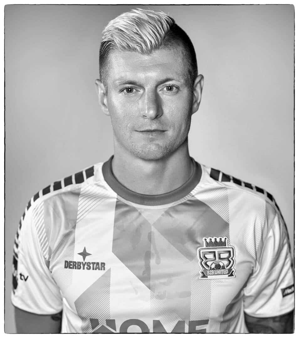 Ein Graustufenbild eines Mannes mit kurzen, hellen Haaren, die nach oben gestylt sind. Er trägt ein gemustertes Fußballtrikot mit Logos, darunter eines, das einer Krone ähnelt, und blickt mit neutralem Gesichtsausdruck in die Kamera. © Fotografie Tomas Rodriguez