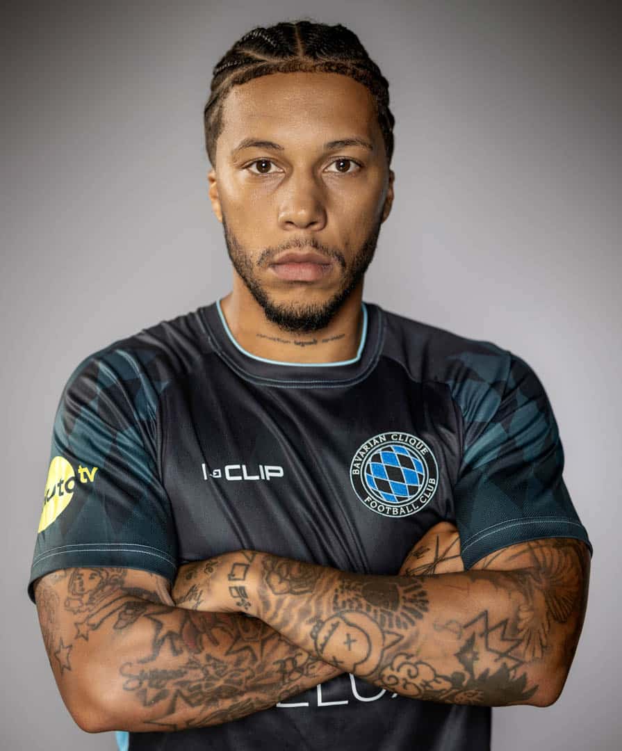 Ein Fußballspieler mit geflochtenen Haaren und Tattoos steht mit verschränkten Armen da. Er trägt ein dunkles Trikot mit einem Logo mit einem Globus auf der Brust und Sponsorenlogos auf den Ärmeln vor einem neutralen Hintergrund. © Fotografie Tomas Rodriguez