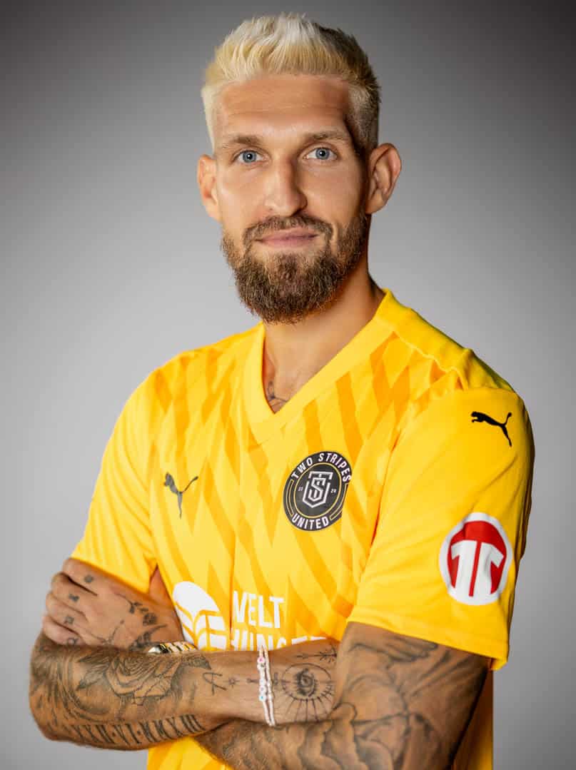 Ein Mann mit Bart und blondem Haar lächelt mit verschränkten Armen und trägt ein leuchtend gelbes Fußballtrikot mit mehreren Logos. Der Hintergrund ist schlicht grau. © Fotografie Tomas Rodriguez