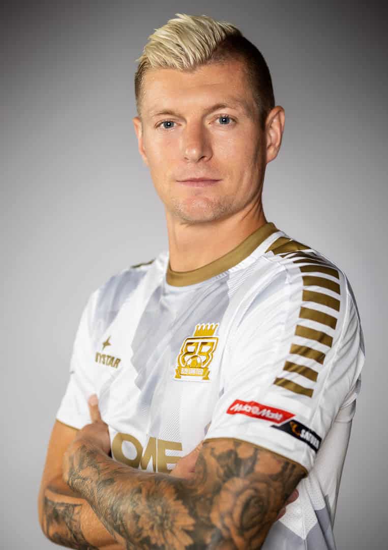 Ein Mann mit kurzen blonden Haaren, der ein weiß-goldenes Sporttrikot trägt, verschränkt die Arme und blickt selbstbewusst in die Kamera. Das Trikot hat Aufnäher und ein Logo auf der Brust. Er hat detaillierte Tattoos auf seinem Unterarm und steht vor einem schlichten grauen Hintergrund. © Fotografie Tomas Rodriguez